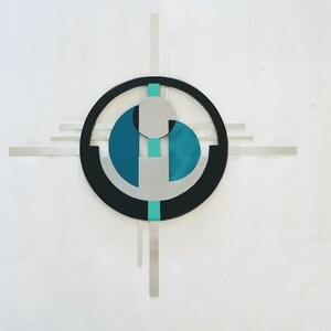 May include: A wall-mounted metal sculpture featuring a central black circle intersected by horizontal and vertical silver bars. The design incorporates teal and silver geometric shapes, including a circular element and two semi-circular forms.