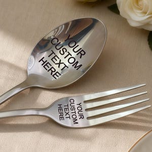 May include: A silver spoon and fork, each engraved with the text "YOUR CUSTOM TEXT HERE". The spoon and fork are crossed on a beige tablecloth. The text "YOUR CUSTOM TEXT HERE" is also on the spoon and fork heads.