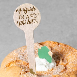 May include: A light brown cake with a wooden topper that reads "A Bride in a Tini bit" with a martini glass illustration. The cake is on a woven plate and has a dollop of white cream and a green decoration.