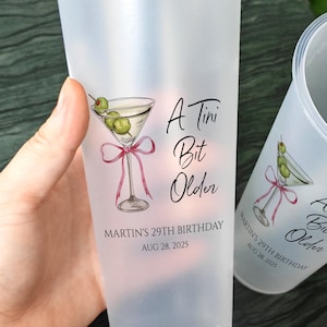 以下が含まれることがあります： マティーニのグラフィックと「A Tini Bit Older」と「MARTIN'S 29TH BIRTHDAY AUG 28, 2025」の文字が入った、2つのフロスト加工されたプラスチックカップ。1つのカップには白いストローが付いています。マティーニのグラフィックには、オリーブとピンクのリボンが含まれています。