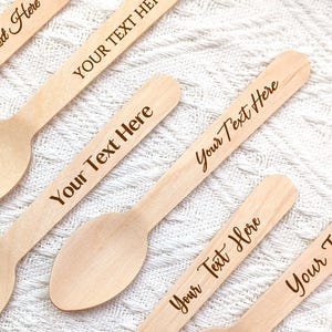 May include: A collection of six light-colored wooden spoons. Each spoon has a rounded bowl and a long handle. The text "Your Text Here" is engraved on each handle in a cursive font. The spoons are arranged on a white, textured surface.