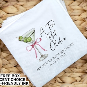 May include: White paper napkins with a martini glass illustration and the text "A Tini Bit Older" with a personalized name and date. The napkins are in a wicker basket.