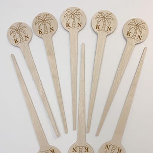 May include: A collection of wooden drink stirrers. Each stirrer features a round top with a laser-etched palm tree design and the initials "K N". The stirrers are light brown in color and have long, slender handles.