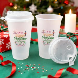 May include: Two clear plastic cups with lids, one assembled and one disassembled, featuring a festive "Nutcracker Season Christmas Mahjong Party" design. The cups are on a green surface with Christmas decorations in the background.
