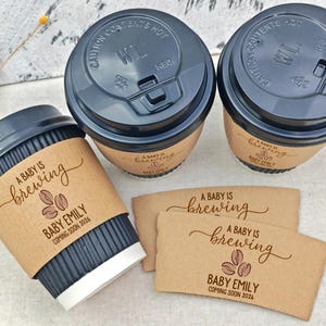 以下が含まれることがあります： 黒い蓋と茶色のスリーブが付いたコーヒーカップが3つ。スリーブには「A Baby Is Brewing」と「Baby Emily」の名前と「2026」の年が書かれています。さらに2つのスリーブも表示されています。