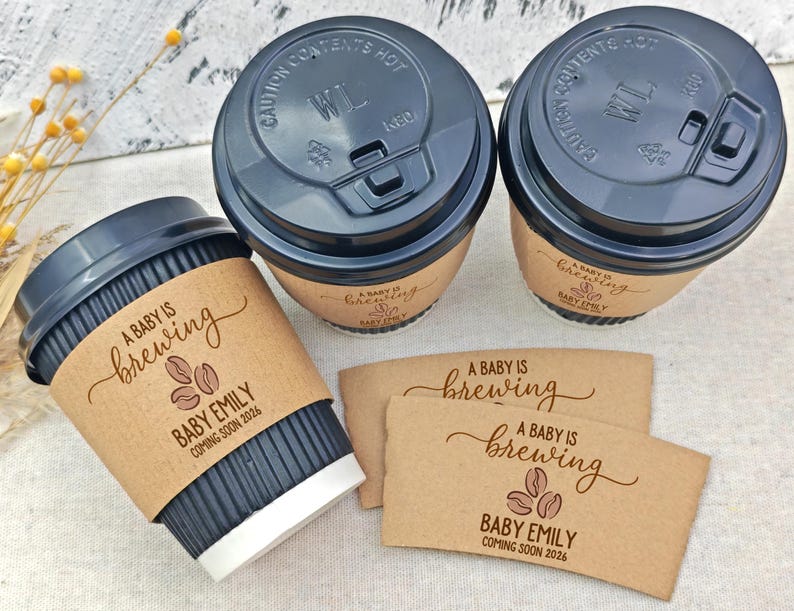 以下が含まれることがあります： 黒い蓋と茶色のスリーブが付いたコーヒーカップが3つ。スリーブには「A Baby Is Brewing」と「Baby Emily」の名前と「2026」の年が書かれています。さらに2つのスリーブも表示されています。