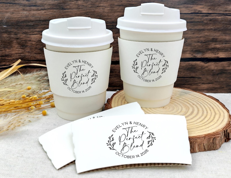 May include: Two off-white paper coffee cups with white lids and matching sleeves. The sleeves are printed with the text "Evelyn & Henry, The Perfect Blend, October 14, 2026" in black script and a floral wreath design. Additional sleeves are visible.
