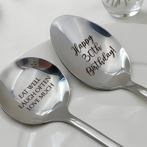 May include: Two shiny silver spoons with engraved text. One spoon reads "Eat Well Laugh Often Love Much." The other spoon says "Happy 30th Birthday!" The spoons are on a white plate.