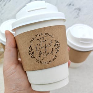 May include: White disposable coffee cups with lids and brown paper sleeves. The sleeves are printed with the text "Evelyn & Henry The Perfect Blend October 14, 2026" in a circular design with leaves. The cups are designed for a wedding or special event.