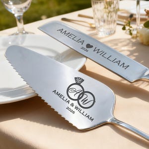 May include: Silver cake server and knife set. The knife is engraved with "AMELIA WILLIAM" and a heart symbol, along with the year "2026". The server features two intertwined rings with the initials "AW" and the same text and year.