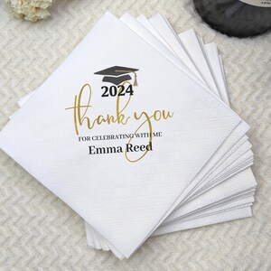 May include: A stack of white paper napkins with a graduation cap graphic and the text "thank you" in gold script. The year "2024" is printed above the text, with the name "Emma Reed" below. The napkins are on a white textured surface.