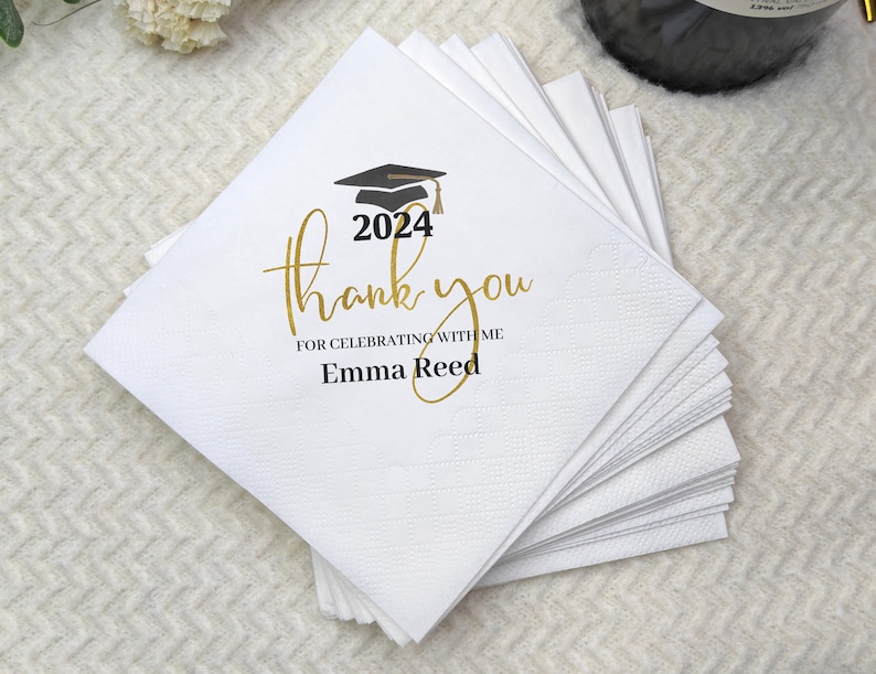 May include: A stack of white paper napkins with a graduation cap graphic and the text "thank you" in gold script. The year "2024" is printed above the text, with the name "Emma Reed" below. The napkins are on a white textured surface.