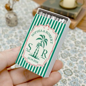 May include: A matchbox with a green and white striped design. The front features a decorative label with the names "Sophia & Robert", a palm tree illustration, and the date 02.14.2026. The matchbox is held in a hand.