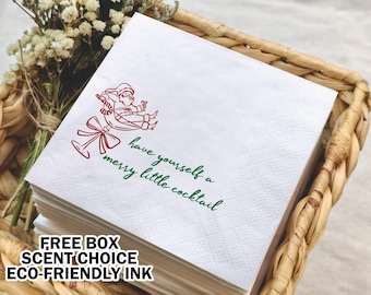 May include: White cocktail napkins with a red illustration of Santa Claus on a cocktail glass, with the text "have yourself a merry little cocktail" in green. The napkins are stacked in a woven basket.