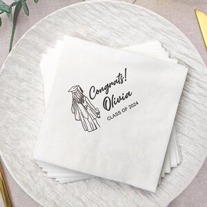 May include: White paper napkins stacked on a white plate. Each napkin is printed with a black illustration of a graduate in a cap and gown, along with the text "Congrats! Olivia CLASS OF 2024."