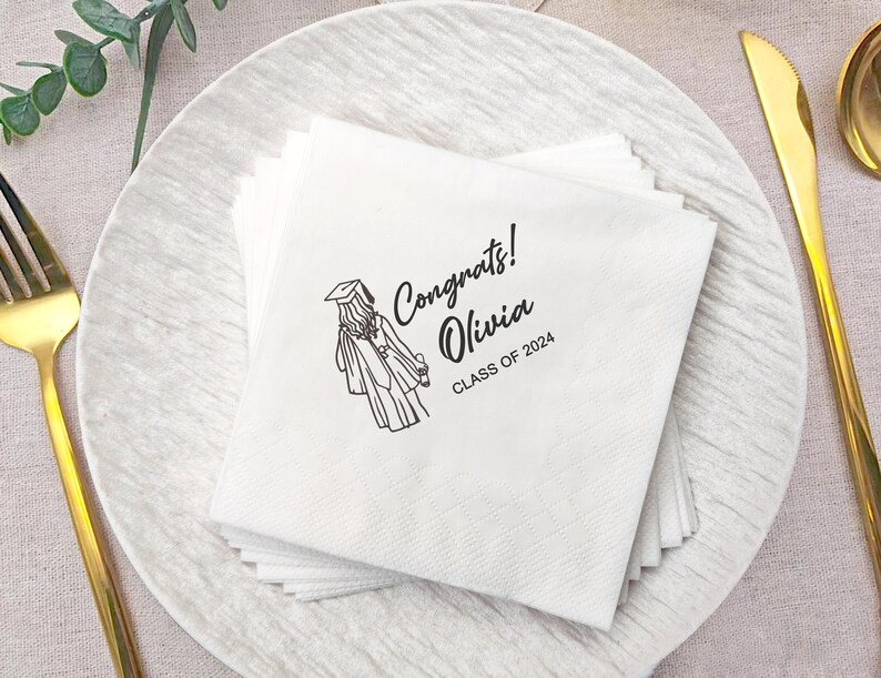 May include: White paper napkins stacked on a white plate. Each napkin is printed with a black illustration of a graduate in a cap and gown, along with the text "Congrats! Olivia CLASS OF 2024."