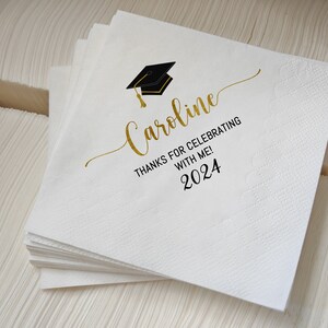 May include: A stack of white paper napkins with a graduation cap graphic and the name "Caroline" in gold script. The text "THANKS FOR CELEBRATING WITH ME! 2024" is also printed on the napkins.