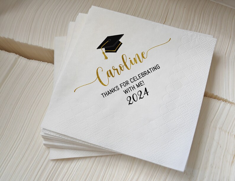 May include: A stack of white paper napkins with a graduation cap graphic and the name "Caroline" in gold script. The text "THANKS FOR CELEBRATING WITH ME! 2024" is also printed on the napkins.