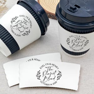 May include: Two coffee cups with black lids and white paper sleeves. The sleeves and cups have the text "Evelyn & Henry The Perfect Blend October 14, 2026" in a circular design with leaves. Two additional sleeves are also visible.
