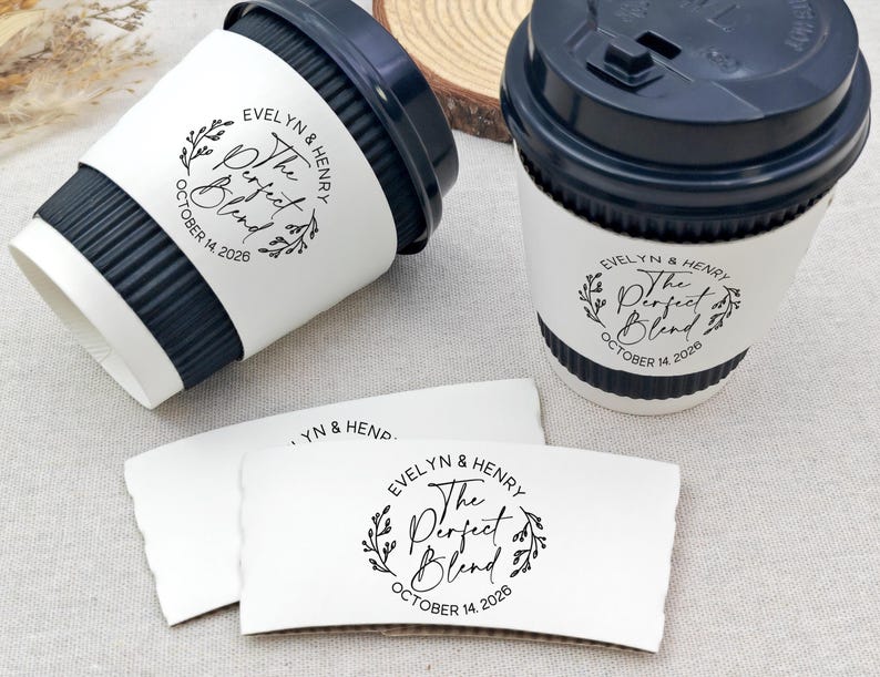 May include: Two coffee cups with black lids and white paper sleeves. The sleeves and cups have the text "Evelyn & Henry The Perfect Blend October 14, 2026" in a circular design with leaves. Two additional sleeves are also visible.