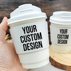 以下が含まれることがあります： 白い使い捨てコーヒーカップが2つ。各カップには、黒で「YOUR CUSTOM DESIGN」と書かれた白いバンドが付いています。片方のカップは手に持たれ、もう片方は木のコースターの上に置かれています。