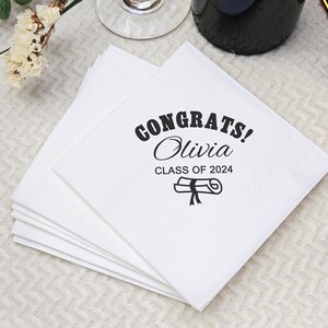 May include: A stack of white paper napkins with the text "CONGRATS! Olivia CLASS OF 2024" printed in black. A graduation cap and scroll graphic is also printed on the napkins. The napkins are square and appear to be made of a textured paper.