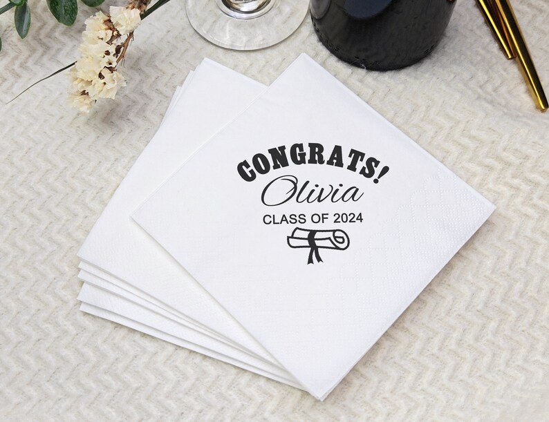 May include: A stack of white paper napkins with the text "CONGRATS! Olivia CLASS OF 2024" printed in black. A graduation cap and scroll graphic is also printed on the napkins. The napkins are square and appear to be made of a textured paper.