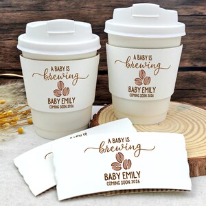 以下が含まれることがあります： 蓋付きの白いコーヒーカップ2つと、お揃いのスリーブ。スリーブには「A Baby is Brewing」とコーヒー豆のグラフィック、「Baby Emily Coming Soon 2026」と書かれています。追加のスリーブも表示されています。