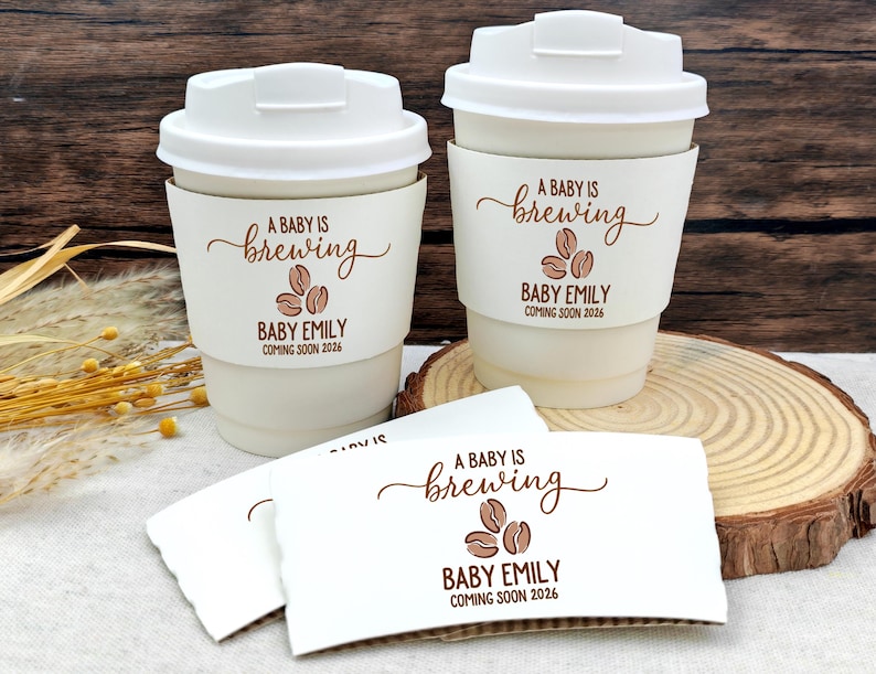 以下が含まれることがあります： 蓋付きの白いコーヒーカップ2つと、お揃いのスリーブ。スリーブには「A Baby is Brewing」とコーヒー豆のグラフィック、「Baby Emily Coming Soon 2026」と書かれています。追加のスリーブも表示されています。