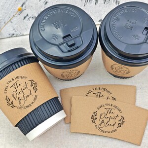 May include: Three coffee cups with black lids and brown sleeves, and two additional sleeves. The sleeves and cups are printed with the text "EVELYN & HENRY The Perfect Blend OCTOBER 14 2026". The cups have a black corrugated exterior.