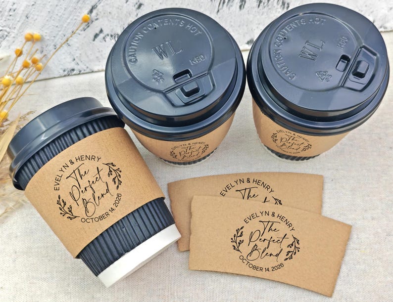 May include: Three coffee cups with black lids and brown sleeves, and two additional sleeves. The sleeves and cups are printed with the text "EVELYN & HENRY The Perfect Blend OCTOBER 14 2026". The cups have a black corrugated exterior.