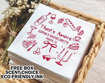 May include: White paper napkins with red ink illustrations of food and drinks, including a pizza slice, wine glass, and a cake. The text "That's Amore" is written in a cursive font, along with "Sophia's Bachelorette Miami, FL".