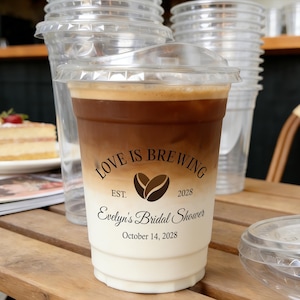 May include: A clear plastic iced coffee cup with a lid, filled with layered coffee and cream. The cup has the text "Love is Brewing" with a coffee bean graphic, "Evelyn's Bridal Shower" and the date October 14, 2028. Other cups are in the background.
