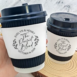 May include: Two coffee cups with navy blue lids and white sleeves. The sleeves are printed with "EVELYN & HENRY The Perfect Blend OCTOBER 14, 2026" in a circular design. One cup is held by a hand.