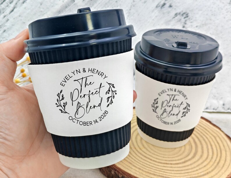 May include: Two coffee cups with navy blue lids and white sleeves. The sleeves are printed with "EVELYN & HENRY The Perfect Blend OCTOBER 14, 2026" in a circular design. One cup is held by a hand.