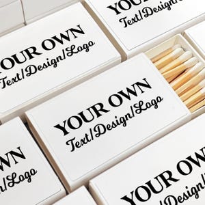 May include: White matchboxes with the text "YOUR OWN Text/Design/Logo" printed in black. One box is open, revealing wooden matches with white tips. The boxes are arranged in a close-up shot.