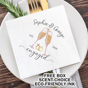 May include: White napkin with black text that reads "Sophie & George" and "EST. 2026 engaged". The napkin features a graphic of two champagne flutes with gold glitter and two gold rings.