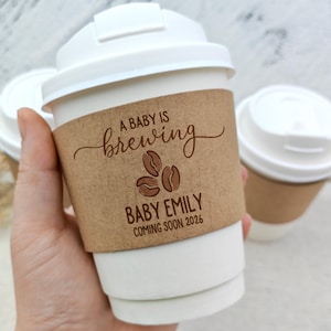 May include: White paper coffee cup with a brown paper sleeve that reads "A BABY IS BREWING" with coffee bean graphics and "BABY EMILY COMING SOON 2026". The cup has a white lid. Another cup is in the background.
