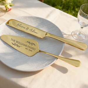 May include: A gold-colored cake knife and server set on a white plate. The knife is engraved with "Olivia & Adam", and the server with "THE WILLIAMS FAMILY EST. 2026". The set is designed for weddings and special occasions.