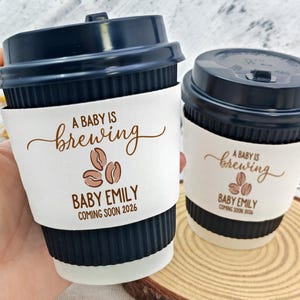 以下が含まれることがあります： ネイビーブルーのコーヒーカップ2つと蓋。各カップには、"A BABY IS brewing"、コーヒー豆、"BABY EMILY"、"COMING SOON 2026"と印刷された白いスリーブが付いています。カップは木製の表面にあります。