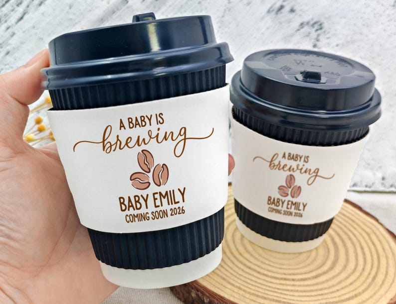 以下が含まれることがあります： ネイビーブルーのコーヒーカップ2つと蓋。各カップには、"A BABY IS brewing"、コーヒー豆、"BABY EMILY"、"COMING SOON 2026"と印刷された白いスリーブが付いています。カップは木製の表面にあります。