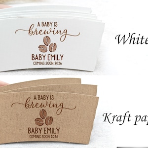 以下が含まれることがあります： 「A BABY IS brewing BABY EMILY COMING SOON 2026」の文字が茶色で印刷された、白とクラフト紙のコーヒーカップスリーブ。スリーブにはコーヒー豆のグラフィックが描かれています。
