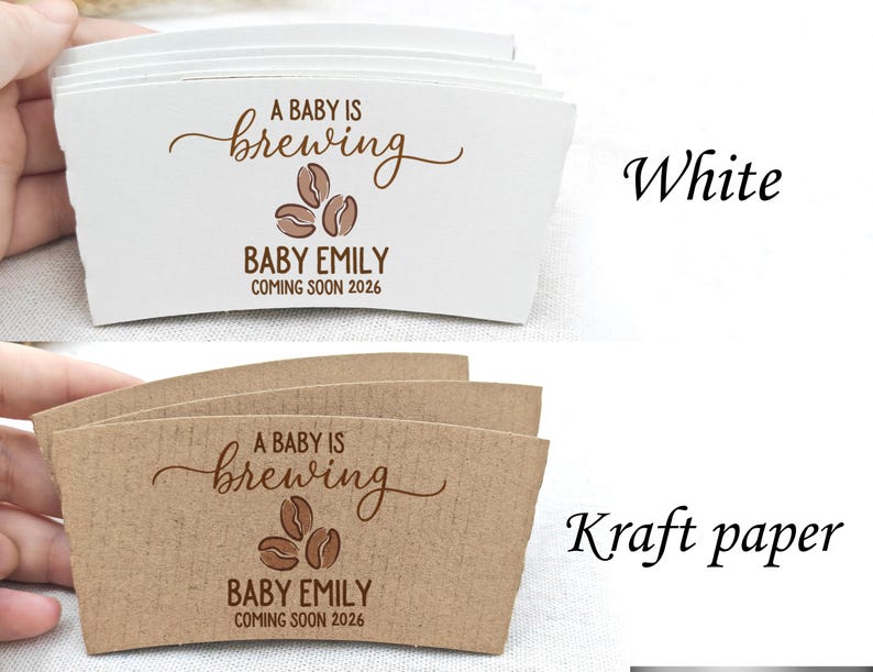 以下が含まれることがあります： 「A BABY IS brewing BABY EMILY COMING SOON 2026」の文字が茶色で印刷された、白とクラフト紙のコーヒーカップスリーブ。スリーブにはコーヒー豆のグラフィックが描かれています。