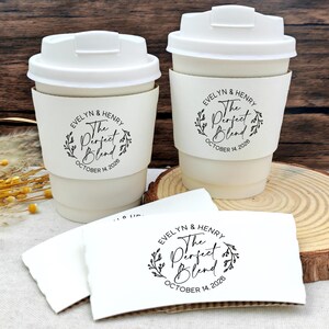 May include: Two off-white paper coffee cups with white lids and matching sleeves. The sleeves are printed with the text "Evelyn & Henry, The Perfect Blend, October 14, 2026" in black script and a floral wreath design. Additional sleeves are visible.