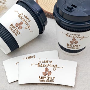 以下が含まれることがあります： 黒い蓋と白いスリーブが付いたコーヒーカップが2つ。スリーブには「A BABY IS brewing」の文字とコーヒー豆のグラフィック、「BABY EMILY COMING SOON 2026」と書かれています。さらに2つのスリーブも表示されています。