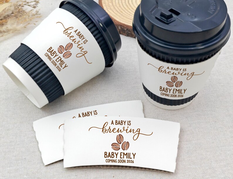 以下が含まれることがあります： 黒い蓋と白いスリーブが付いたコーヒーカップが2つ。スリーブには「A BABY IS brewing」の文字とコーヒー豆のグラフィック、「BABY EMILY COMING SOON 2026」と書かれています。さらに2つのスリーブも表示されています。