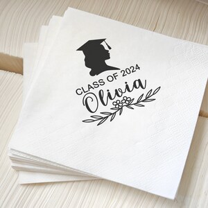 May include: A stack of white paper napkins with a graduation cap silhouette and the text "Class of 2024 Olivia" in black. The napkins have a subtle textured pattern and are likely for a graduation celebration.