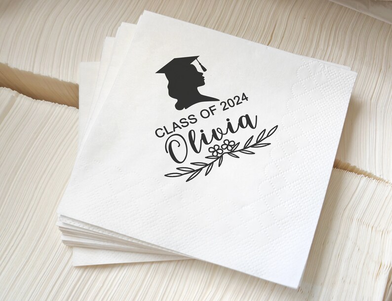 May include: A stack of white paper napkins with a graduation cap silhouette and the text "Class of 2024 Olivia" in black. The napkins have a subtle textured pattern and are likely for a graduation celebration.