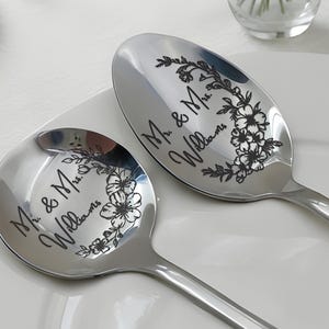 Può includere: Due cucchiai d'argento da portata con disegni floreali incisi e la scritta "Mr. & Mrs. Williams". I cucchiai sono su un piatto bianco, ideali per un regalo di nozze o anniversario. Superficie lucida e riflettente.