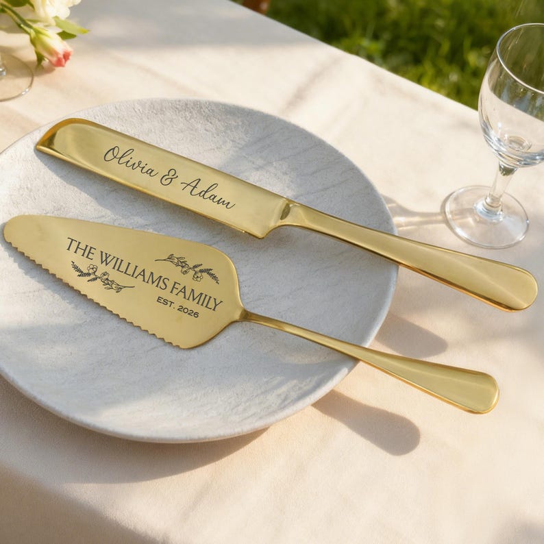 May include: A gold-colored cake knife and server set on a white plate. The knife is engraved with "Olivia & Adam", and the server with "THE WILLIAMS FAMILY EST. 2026". The set is designed for weddings and special occasions.